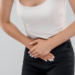 Clolse up of bending incognito caucasian woman in casual suit posing over gray isolated background and touching lower abdomen because of ache. Front crop of brunette suffering from pain in stomach.